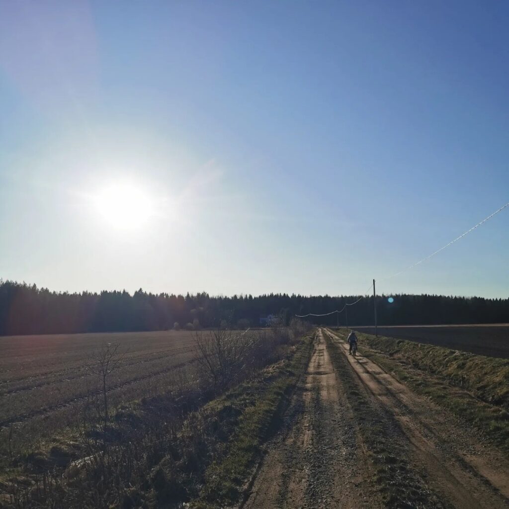 Kuja talolle peltojen keskellä ruskeassa maisemassa, jonka taustalla sininen taivas ja aurinko.